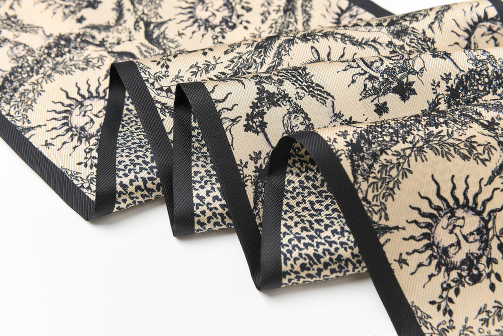 A luxurious 16 Momme silk twill long scarf from SusanSilk, showcasing an intricate sun and floral pattern in beige and black, elegantly bordered by black trim.