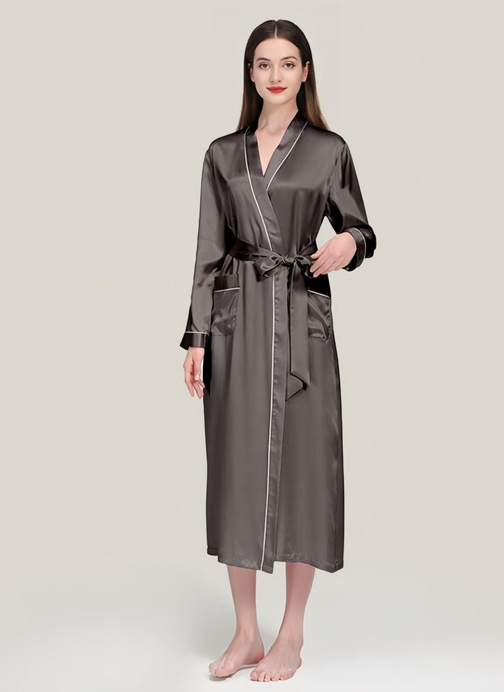 A woman wearing the SusanSilk 22 Momme Contrast Piping Long Silk Robe for women, featuring two front pockets and a waist tie, stands barefoot against a plain background.