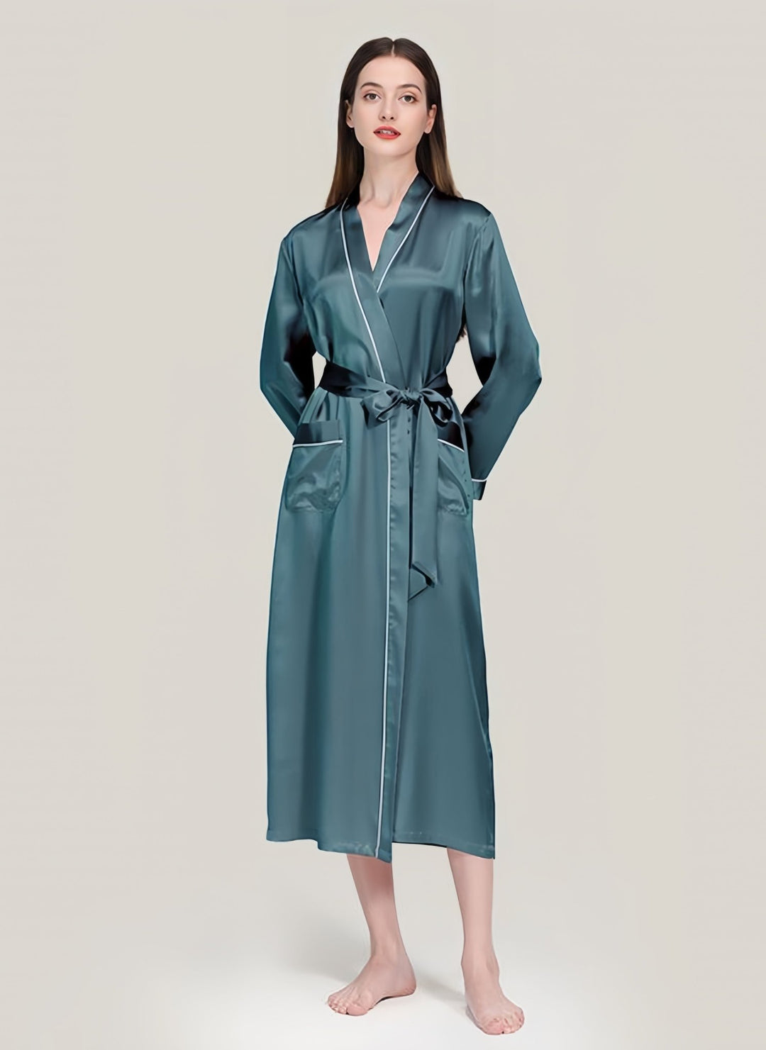 A person stands barefoot, wrapped in a long 22 Momme Contrast Piping Silk Robe from SusanSilk, featuring a belt and two pockets, against a plain light background—the scene exudes an air of quiet elegance reminiscent of fine susansilk.