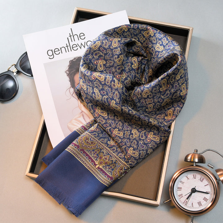 A Paisley Men's Silk Twill Scarf - Navy from SusanSilk is draped over an open book titled "The Gentleman" on a table. Silk sunglasses, an alarm clock, and a framed picture are also on the table.