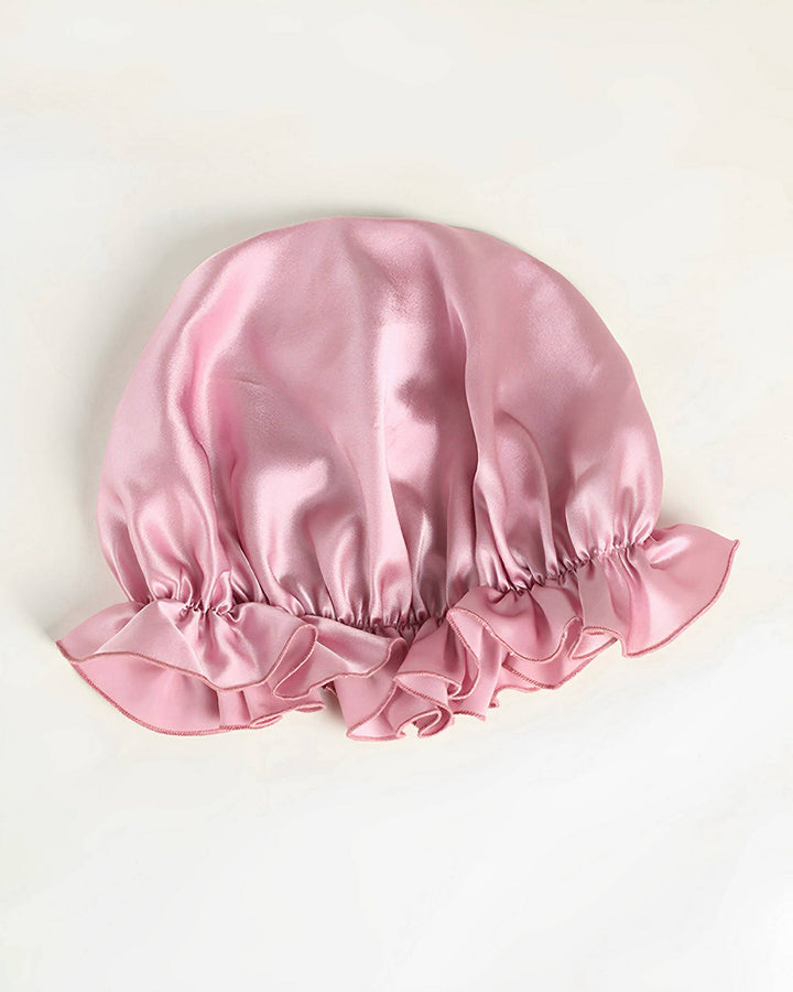 The Flounced Silk Bonnet For Sleeping by SusanSilk, featuring pink satin with ruffled edges, is displayed on a white background.