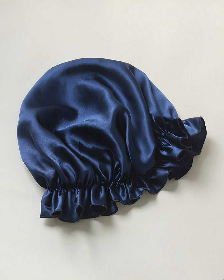 A navy blue Flounced Silk Bonnet For Sleeping by SusanSilk, featuring ruffled edges, is displayed against a plain light background.