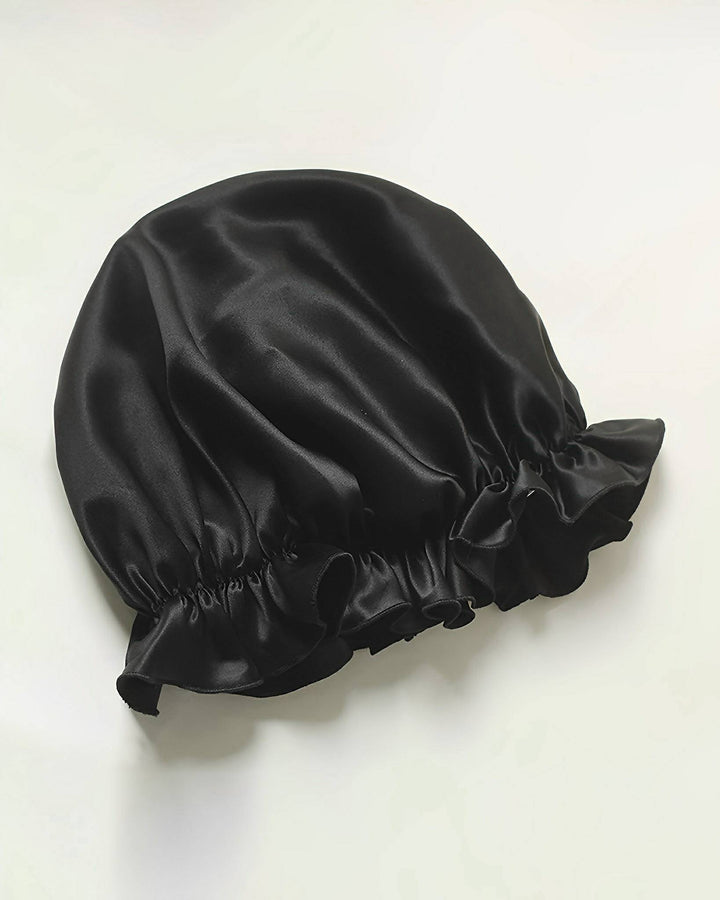 A Flounced Silk Bonnet For Sleeping from SusanSilk, featuring a black satin finish and an elasticated edge, lays flat against a white background.