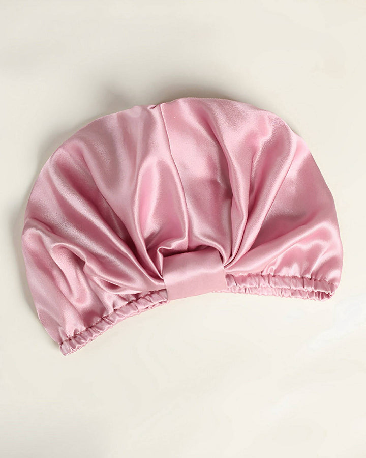 An Elegant Pleated Silk Bonnet For Sleeping from SusanSilk, featuring a light pink hue and an elastic band, lies flat on a cream-colored surface.