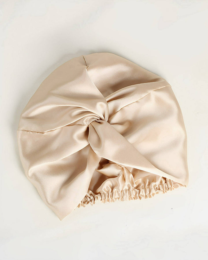 The SusanSilk Luxury Turban Silk Sleep Cap is a beige satin bonnet crafted from luxurious susansilk, featuring a knot design at the front and gathered elastic at the back.