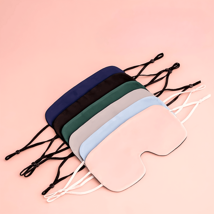 An array of six SusanSilk Oversized Silk Eye Masks in different colors (pink, blue, green, gray, black, and navy) are laid out on a peach-colored surface. Each luxurious mask has adjustable straps.