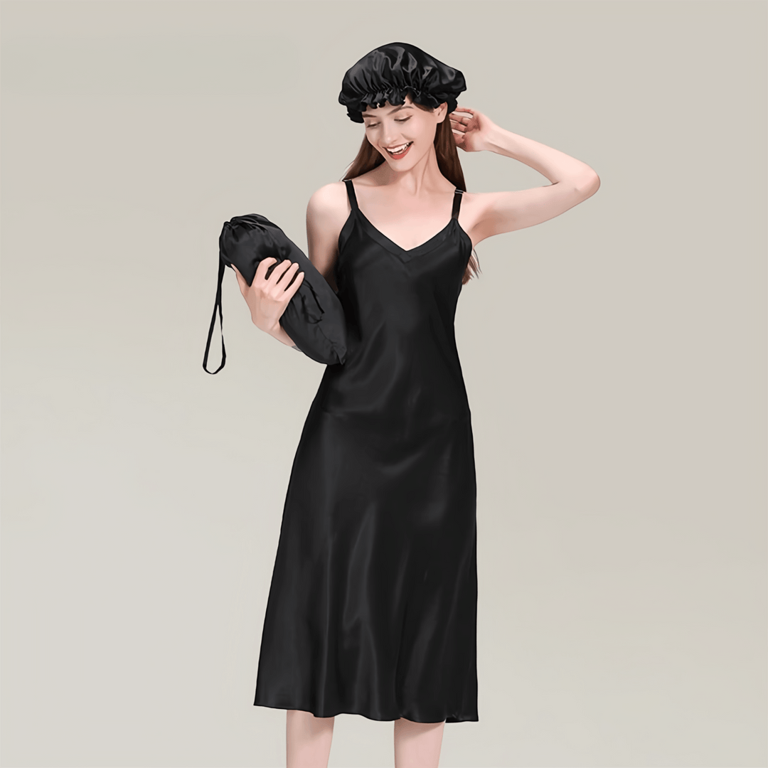 A woman wearing a 19 Momme Midi Silk Nightdress by SusanSilk and a black hair bonnet is standing, smiling, and holding a black drawstring bag against a plain background.