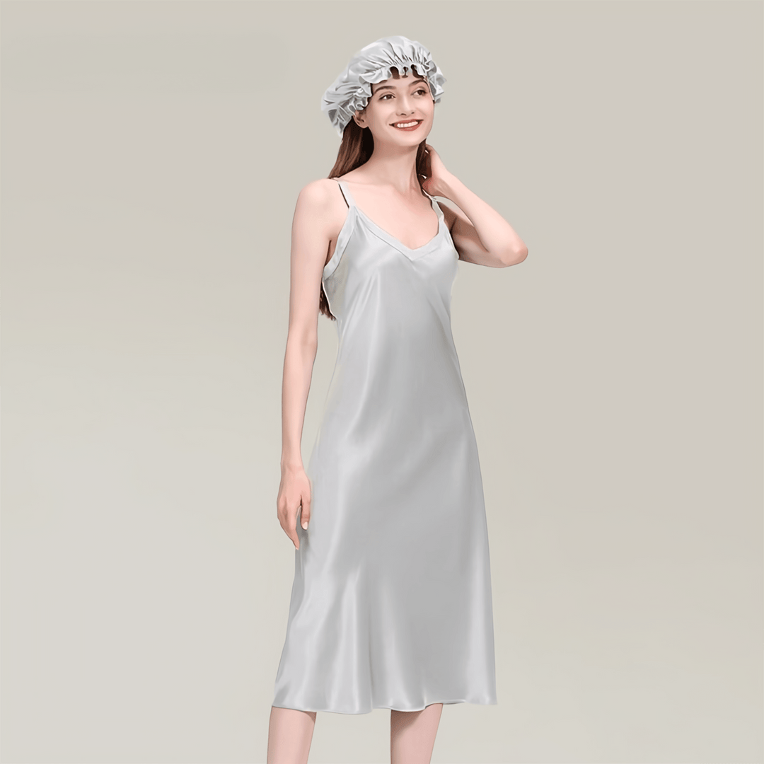 A woman dressed in a light-colored, sleeveless 19 Momme Midi Silk Nightdress from SusanSilk and a matching hair cap is posing against a plain background. She is smiling and has her hand on her neck.