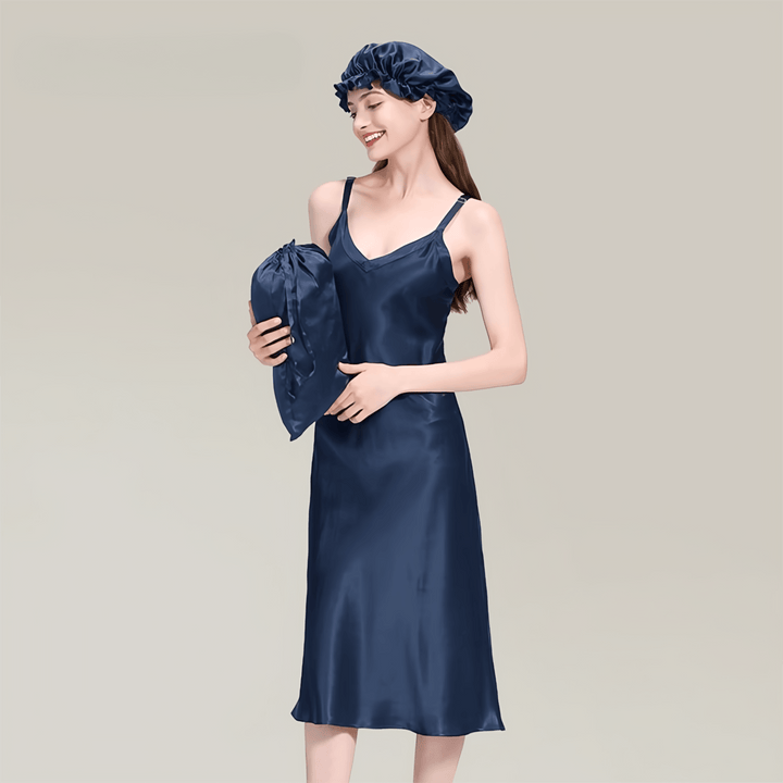 A woman wearing a SusanSilk 19 Momme Midi Silk Nightdress and a matching bonnet holds a silk pillowcase against a plain background.