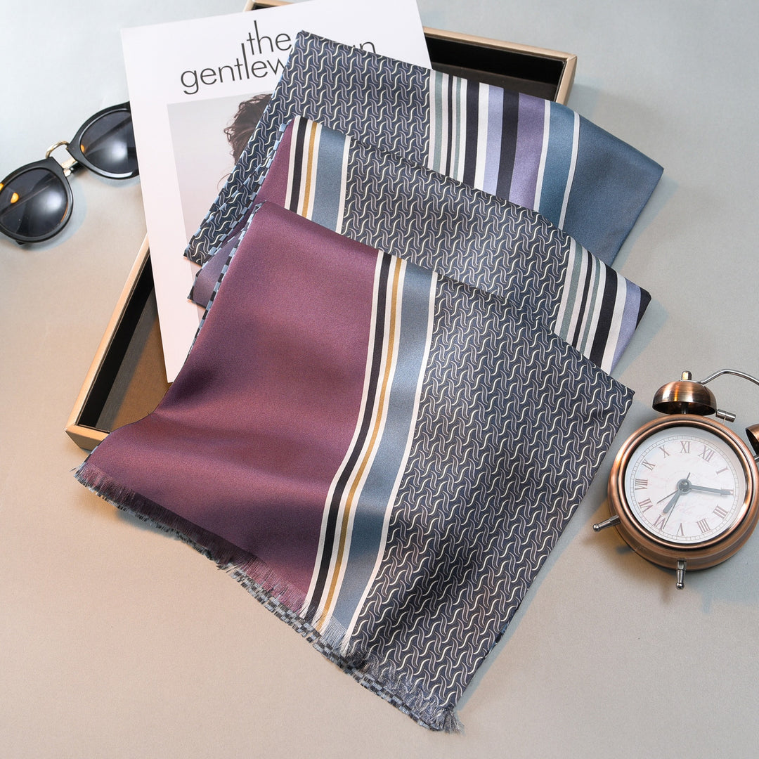 A SusanSilk Luxurious Men's Silk Twill Scarf in Grey Khaki is neatly arranged on a tray beside a pair of sunglasses and a magazine featuring a woman. An alarm clock is positioned at the bottom right corner of the tray.