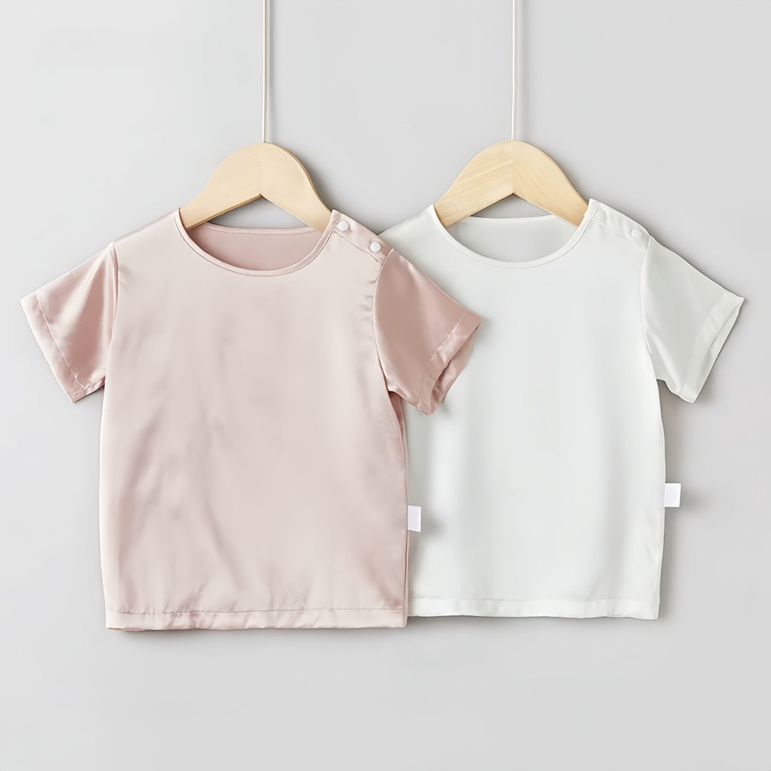 Two short-sleeve tops, one pink and one white, made from luxurious 19 Momme Mulberry Silk from SusanSilk's Baby & Toddler line, are displayed on wooden hangers against a plain gray background.