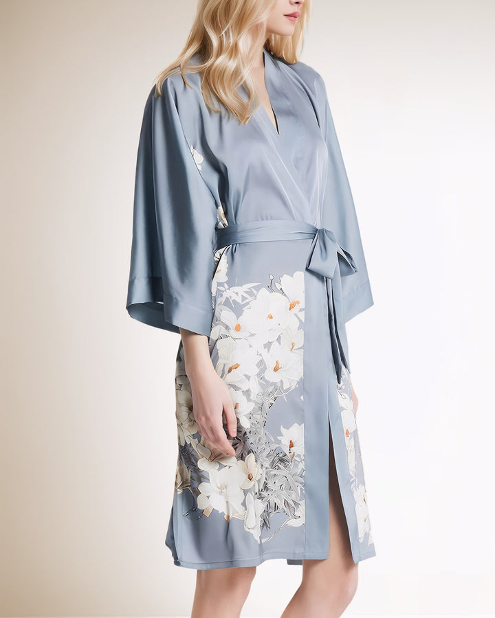 A person wearing the White Lilies Short Silk Kimono by SusanSilk, featuring a light blue, floral pattern, stands against a plain background.