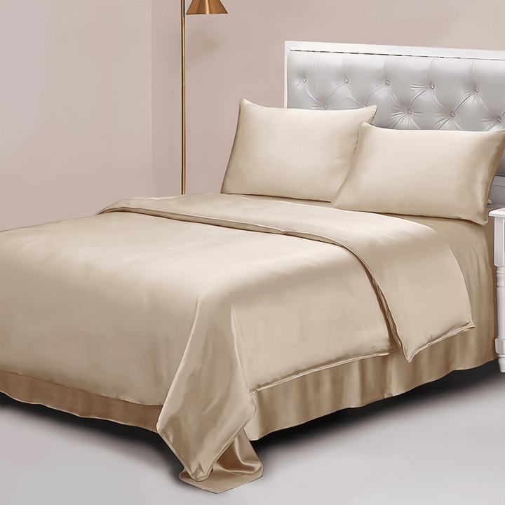 A neatly made bed adorned with the 22 Momme Luxury Silk Bedding Set - 4Pcs by SusanSilk, featuring beige satin sheets, two plush pillows, and a luxurious silk comforter. A tufted headboard and a nightstand with a lamp are visible in the background.