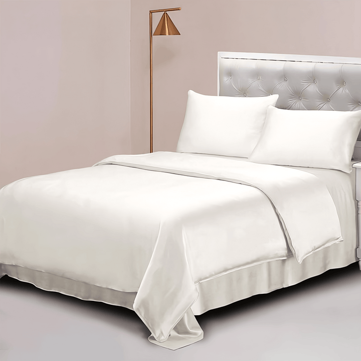 A neatly made bed with white silk linens from the SusanSilk 22 Momme Luxury Silk Bedding Set - 4Pcs, two white pillows, and a tufted headboard. A bedside table with a lamp is on the right side. A standing lamp with a gold finish is on the left side.