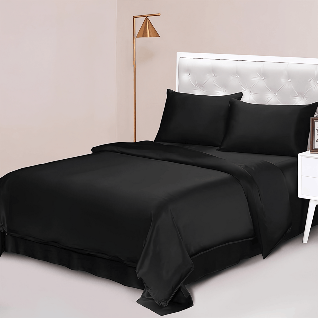 A neatly made bed with SusanSilk's 22 Momme Luxury Silk Bedding Set - 4Pcs in black, two pillows, and a tufted white headboard. A modern floor lamp and nightstand with a picture frame are beside the bed.