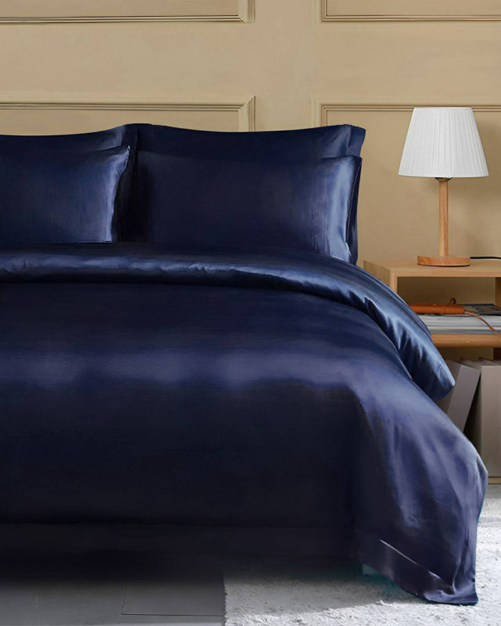 A neatly made bed adorned with a dark blue SusanSilk 22 Momme Seamless Silk Duvet Cover with Flange Border, two pillows, and a bedside table featuring a lamp in a lightly colored room.