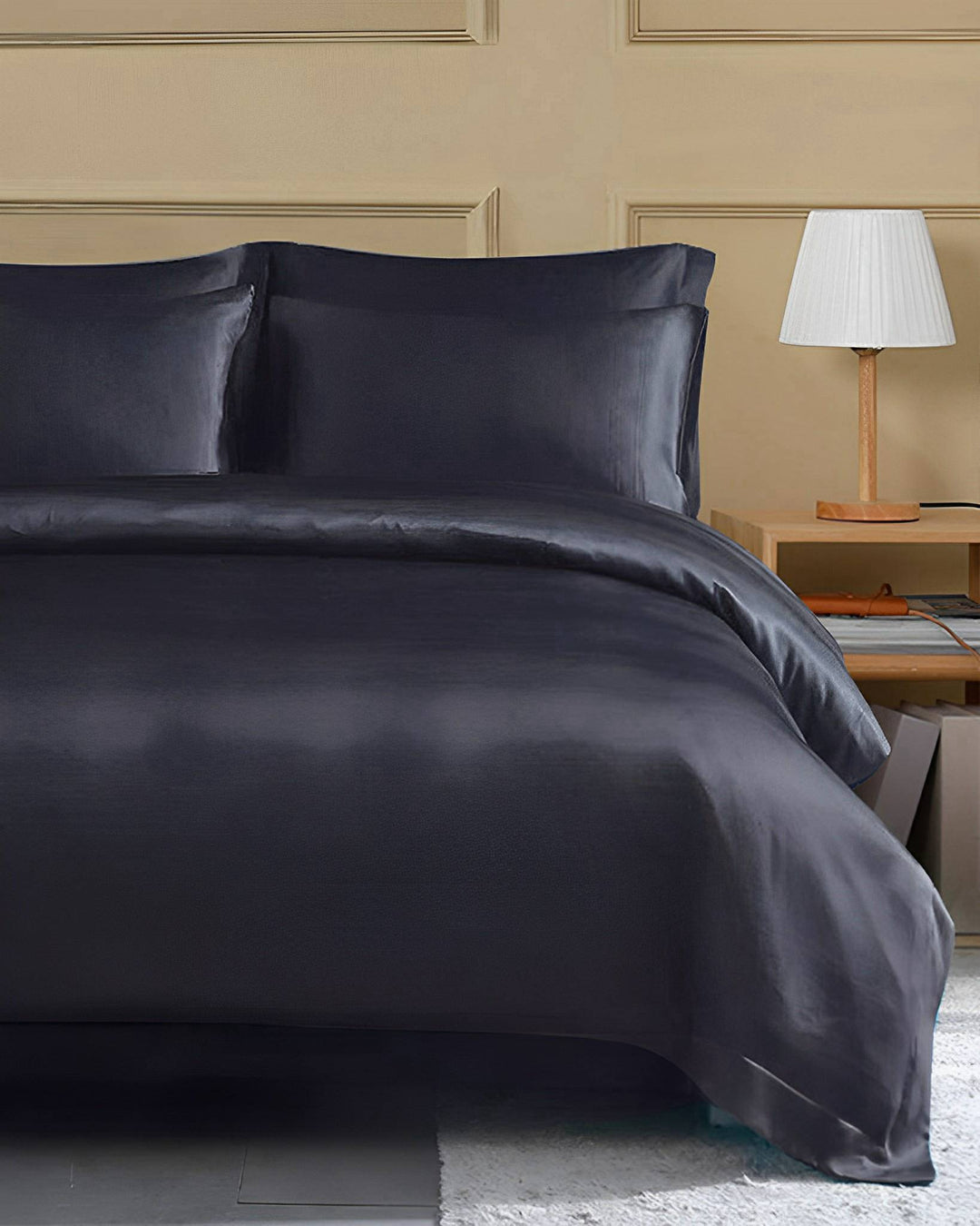 A neatly made bed with SusanSilk's 22 Momme Seamless Silk Duvet Cover in dark grey, flange border, including pillows and a comforter, stands against a beige paneled wall. A bedside table with a white lampshade is on the right.