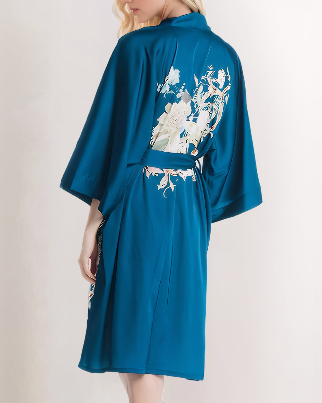 A person with long blonde hair, wearing a blue Peony Floral Short Silk Kimono from SusanSilk, stands facing away. The delicate fabric of the robe catches the light, adding an extra touch of elegance to its floral embroidery on the back.