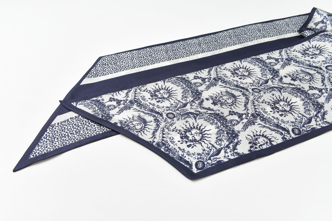 A "Gerbera" scarf by SusanSilk, featuring intricate blue and white patterns with a blue border, is crafted from affordable 16 Momme luxury silk twill and displayed flat on a white surface.