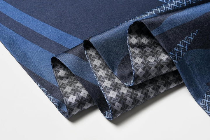 Close-up of a folded silk fabric in Dreamy Blue hues. The top section of the Luxurious Men's Silk Twill Scarf by SusanSilk features a solid dark blue and striped design, while the underside showcases an intricate geometric pattern in shades of gray, capturing exquisite craftsmanship.