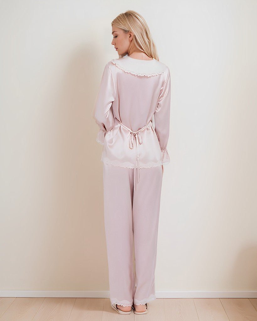 A woman stands facing away, wearing SusanSilk's 22 Momme Lace Silk Pajamas Set in light pink, made of luxurious silk with delicate lace trim, standing in a neutral-colored room.