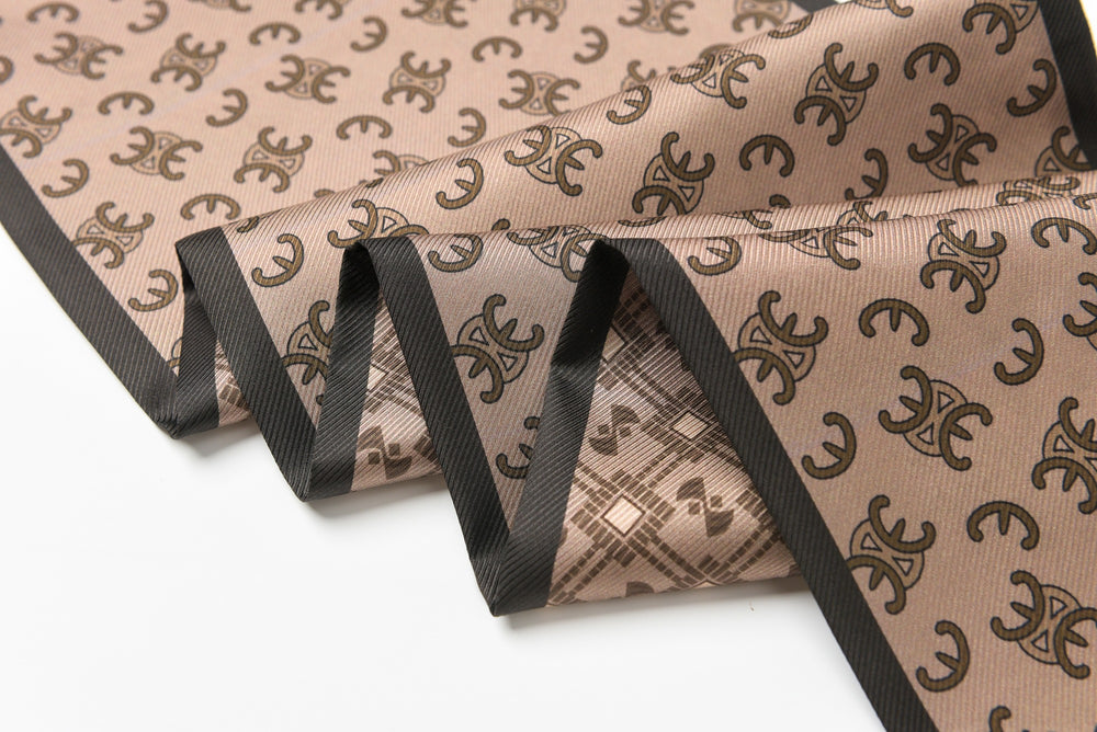 Close-up of the 16 Momme Affordable Luxury Silk Twill Long Silk Scarf-Classic Arc de Triomphe by SusanSilk, showcasing its beige silk fabric adorned with a black pattern of geometric shapes and symbols, folded in a zigzag manner with black trimming along the edges.