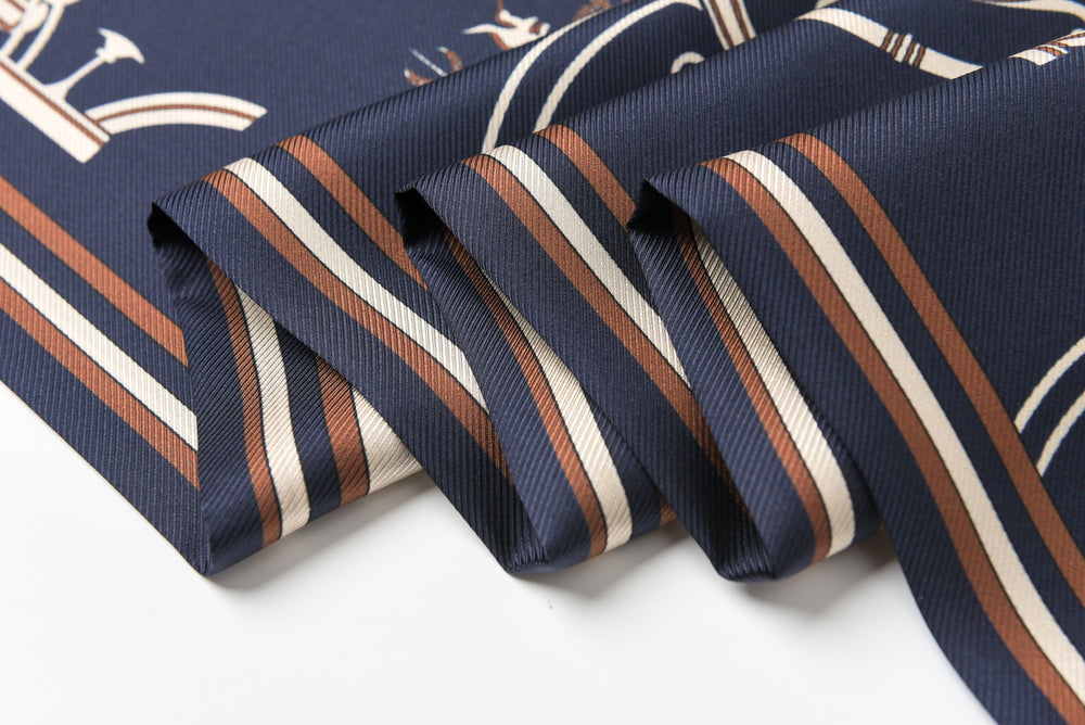 A close-up of the 16 Momme Affordable Luxury Silk Twill Long Silk Scarf-Blue Striped Wagon by SusanSilk, showcasing its folded navy blue silk fabric with beige and brown striped patterns.