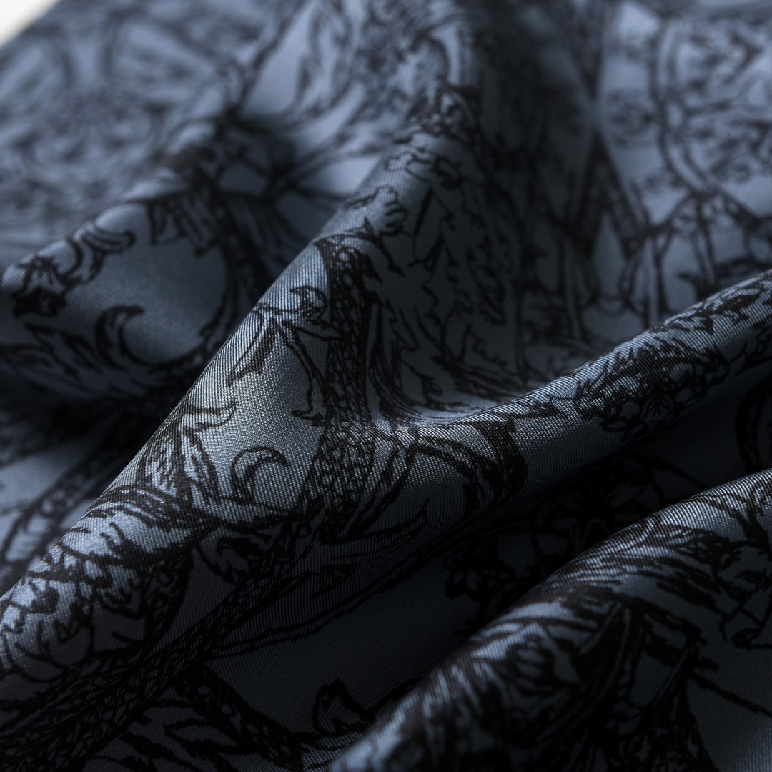 Close-up of the Luxurious Men's Silk Twill Scarf - Gentleman Gray by SusanSilk, featuring crumpled silk fabric with black, intricate floral patterns on a dark gray background. The texture of the material appears smooth and slightly reflective.