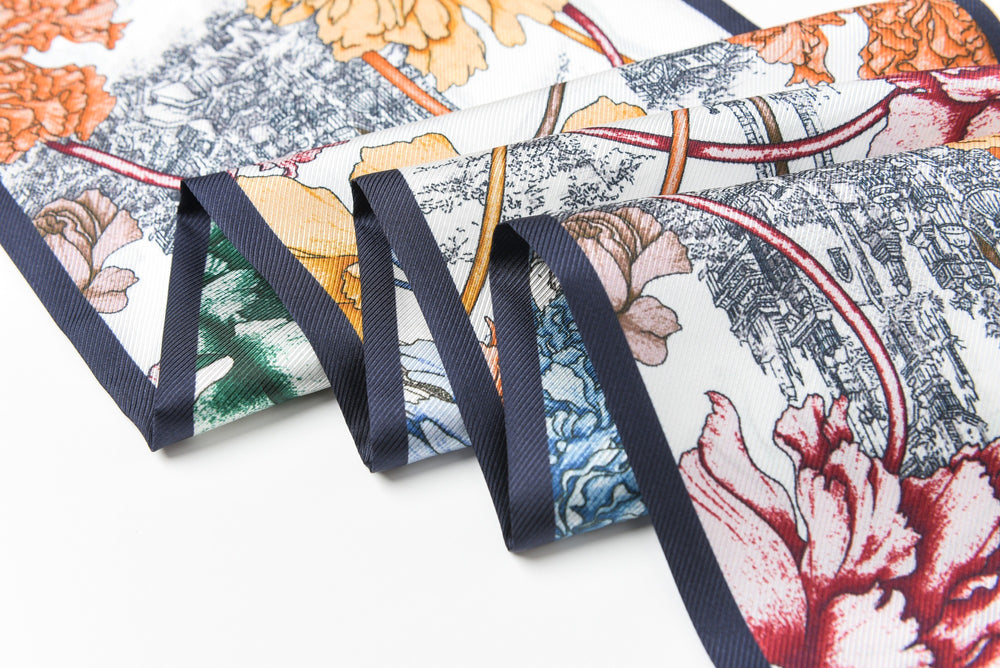 A close-up view of the 16 Momme Affordable Luxury Silk Twill Long Silk Scarf-Peony Mirage from SusanSilk, showcasing its folded, floral-printed silk fabric in various colors with detailed illustrations and a dark border.