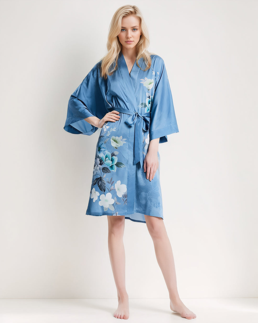 A person with long blonde hair is wearing a Blue Floral Short Silk Kimono from SusanSilk, standing barefoot against a plain white background.