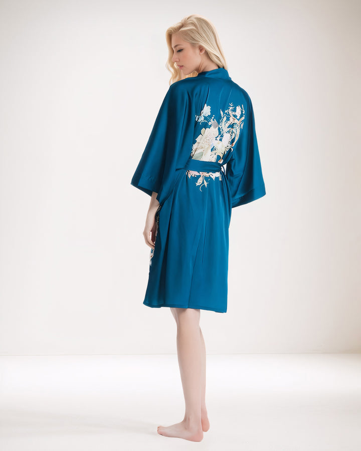 A person with long blonde hair stands barefoot in a blue Peony Floral Short Silk Kimono by SusanSilk, featuring an intricate white design on the back, facing away from the camera.