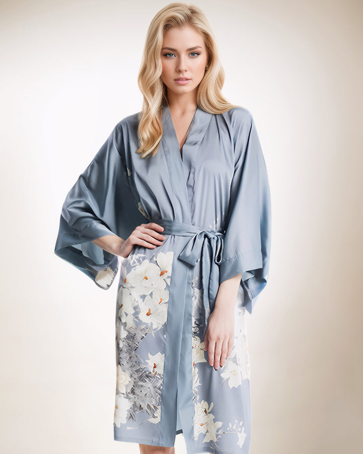 A woman with long blonde hair wears the White Lilies Short Silk Kimono by SusanSilk, a light blue silk robe adorned with white floral patterns. She stands against a neutral background.