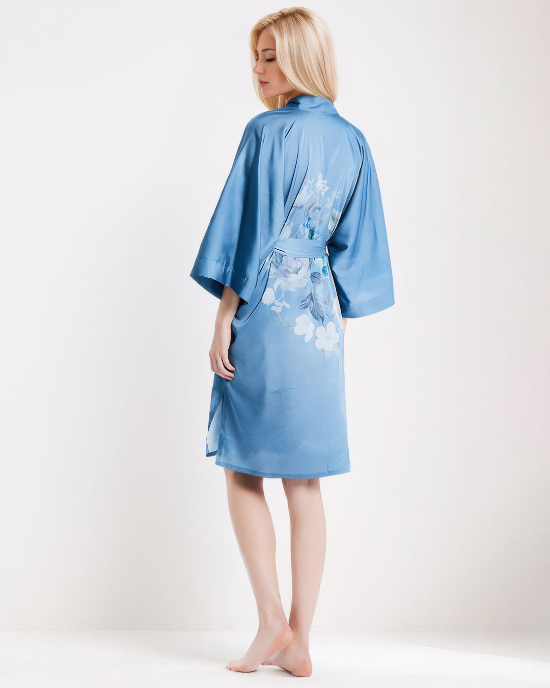 A person stands barefoot in a Blue Floral Short Silk Kimono by SusanSilk, facing slightly away from the camera in a well-lit room.