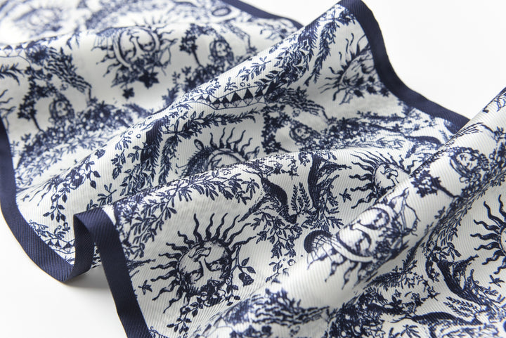 Close-up of a folded SusanSilk 16 Momme Affordable Luxury Silk Twill Long Silk Scarf-Gerbera, showcasing a detailed blue and white pattern with intricate designs, including sun and floral motifs.