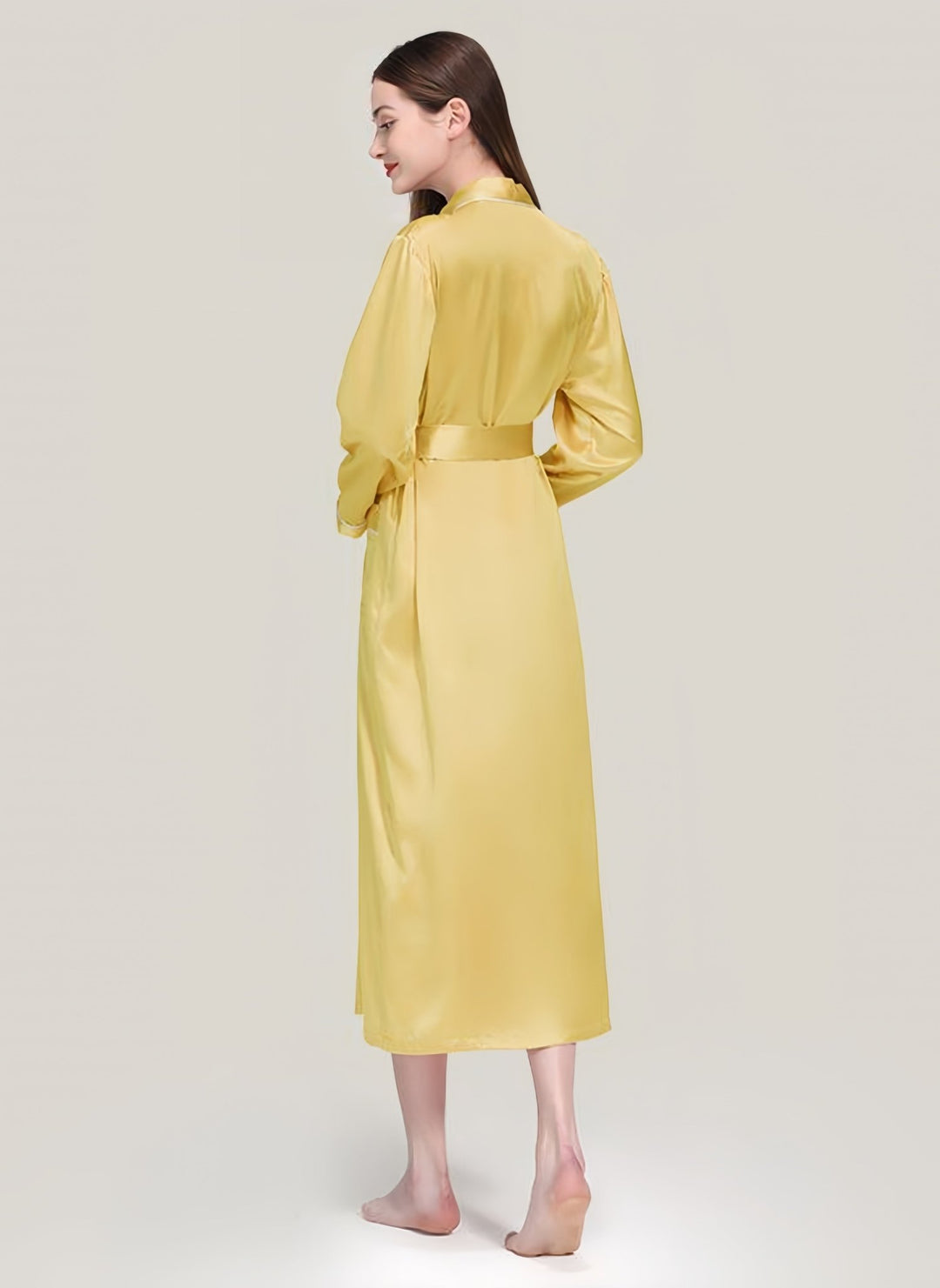 A woman in the luxurious SusanSilk 22 Momme Contrast Piping Long Silk Robe, crafted from yellow satin, stands barefoot with her back to the camera against a plain, light-colored background.