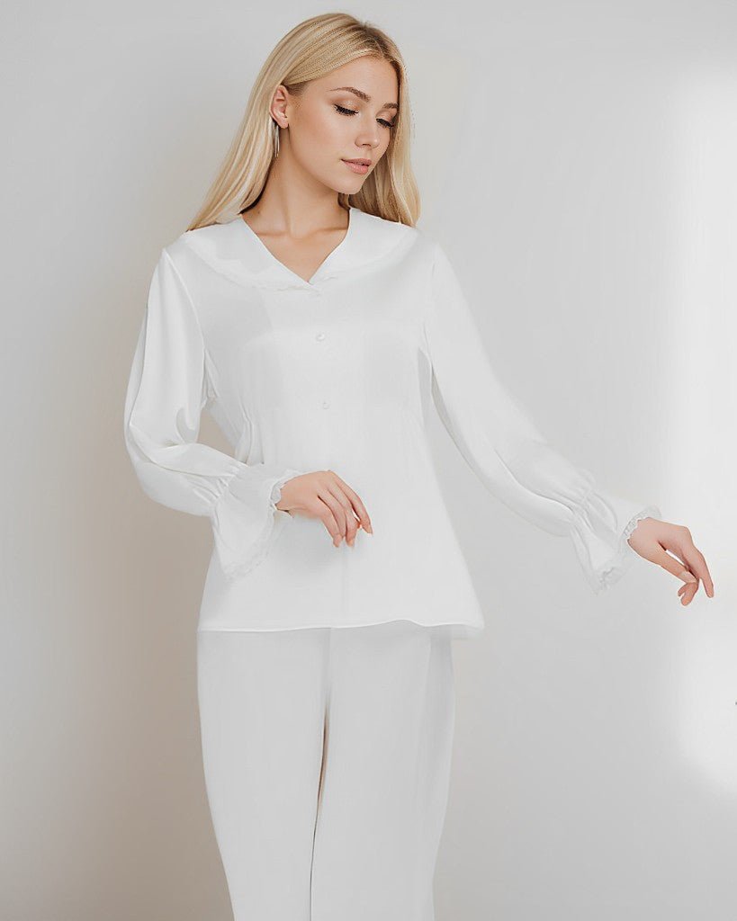 A woman with long blonde hair is standing against a plain background, looking down and touching her blouse with one hand. She is wearing the 22 Momme Lace Silk Pajamas Set by SusanSilk, which consists of a luxurious white silk blouse and matching white pants.