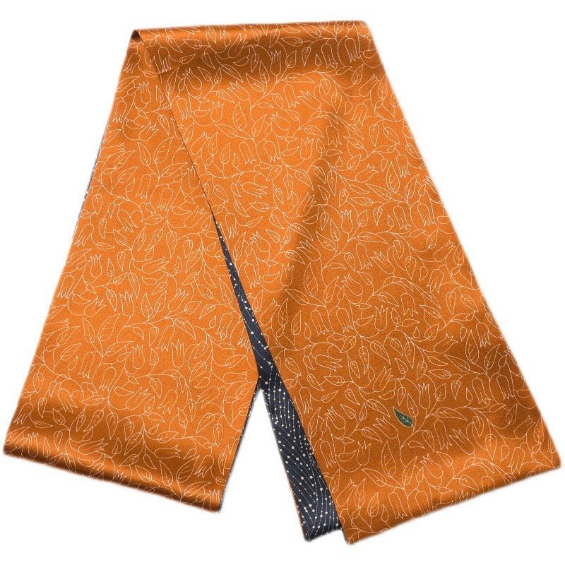 An Orange Flower Sprigs Silk Long Fluttering Belt from SusanSilk, showcasing an orange silk fabric adorned with a floral pattern and a small green leaf logo. The reverse side highlights a dark blue design with white dots.