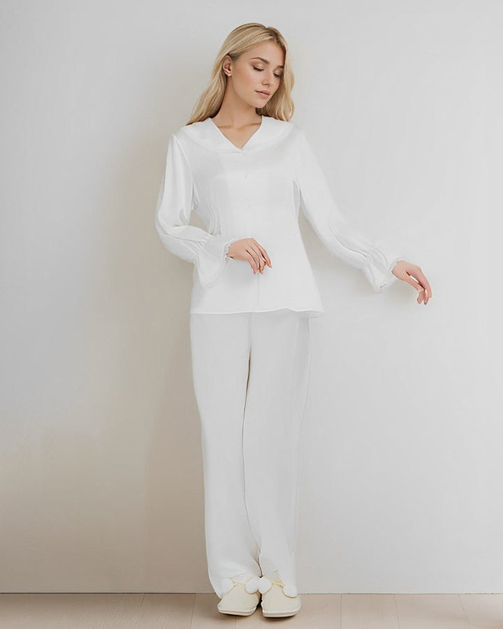 A woman with long blonde hair is standing against a plain white wall, dressed in an elegant white SusanSilk 22 Momme Lace Silk Pajamas Set, white trousers, and matching shoes. She is looking down with her hands relaxed at her sides.