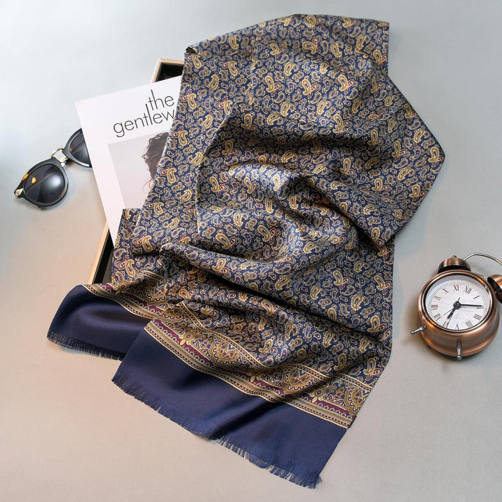 A SusanSilk Paisley Men's Silk Twill Scarf in Navy is elegantly laid over a magazine, with sunglasses and a pocket watch placed nearby on a flat surface.