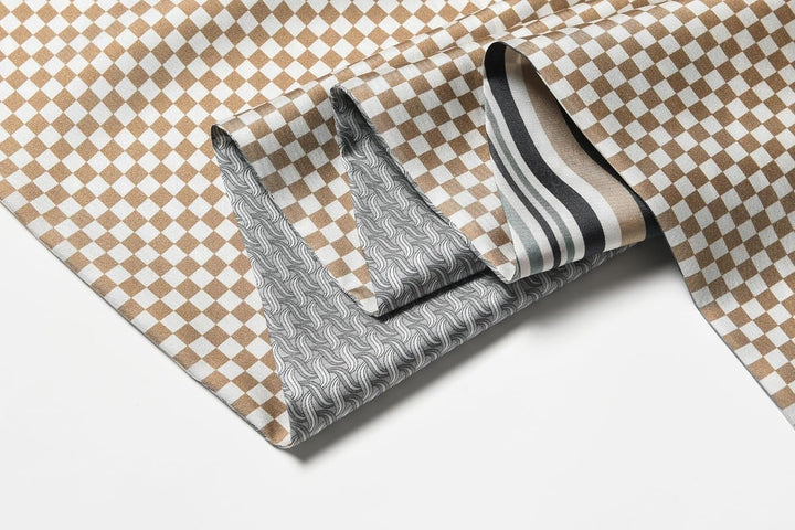 A folded Luxurious Men's Silk Twill Scarf in Grey Khaki from SusanSilk showcases a brown and white checkered pattern on one side, while its underside reveals two distinct patterns: wavy lines and stripes in shades of gray and beige.