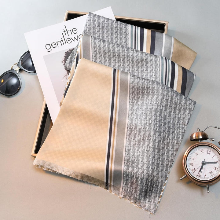 A Luxurious Men's Silk Twill Scarf in Grey Khaki by SusanSilk is draped over an open box containing a magazine titled "The Gentlewoman." Nearby are sunglasses and a brass alarm clock.