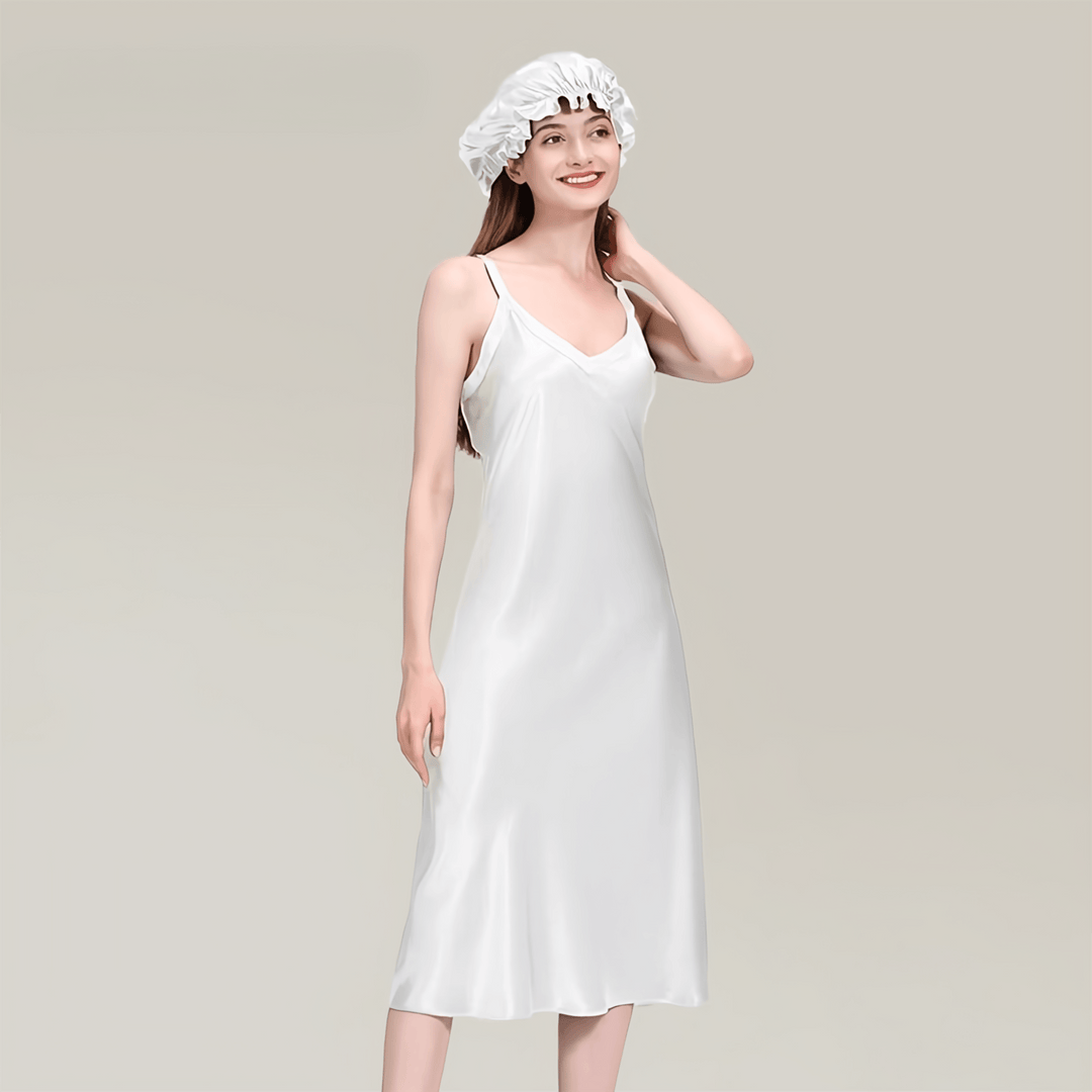 A woman in a 19 Momme Midi Silk Nightdress from SusanSilk and a matching hair bonnet stands against a neutral background. She is smiling and touching her hair.