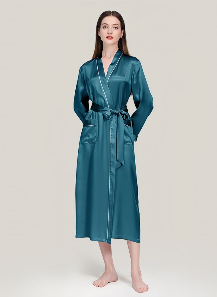 A woman stands barefoot against a plain background, wearing the 22 Momme Contrast Piping Long Silk Robe from SusanSilk. The robe's long, teal satin fabric with pockets and a tied belt showcases a luxurious sheen, adding an extra touch of elegance to her look.