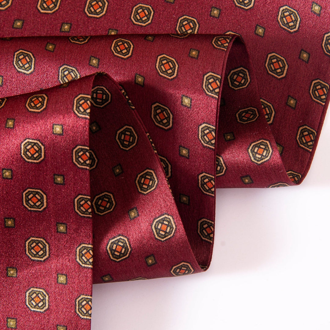 A close-up of a folded Geometric Pattern Long Silk Scarf by SusanSilk, featuring small, square motifs in black and gold on luxurious red fabric.