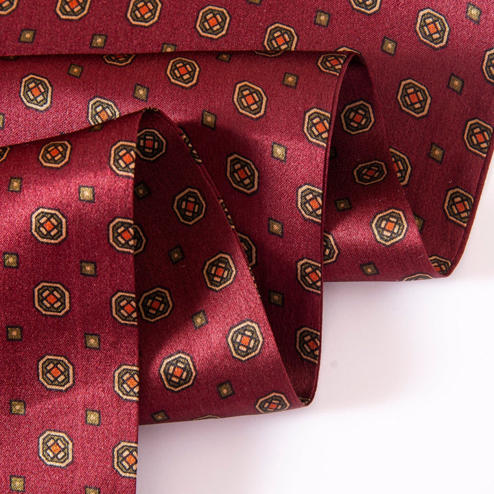 A close-up of a folded Geometric Pattern Long Silk Scarf by SusanSilk, featuring small, square motifs in black and gold on luxurious red fabric.