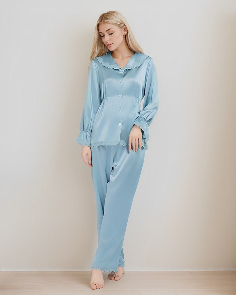 A woman is posed against a plain wall, clad in SusanSilk's 22 Momme Lace Silk Pajamas Set in a light blue hue. The luxurious silk shimmers gently as it catches the light. Her relaxed demeanor is evident as she looks down, with her long hair cascading freely around her shoulders.