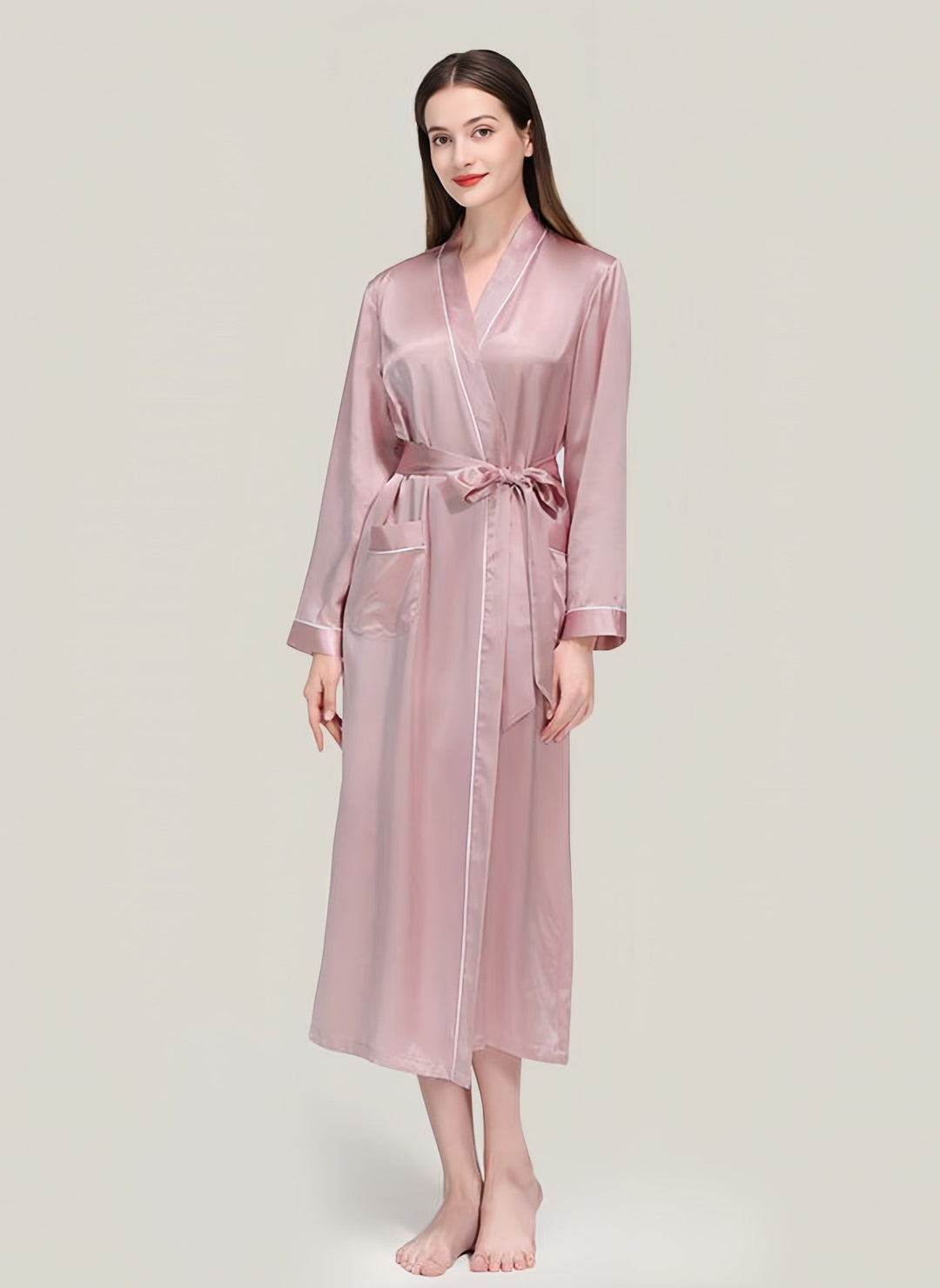 A barefoot woman stands against a plain background, wearing the 22 Momme Contrast Piping Long Silk Robe for Women by SusanSilk, in light pink and tied at the waist.