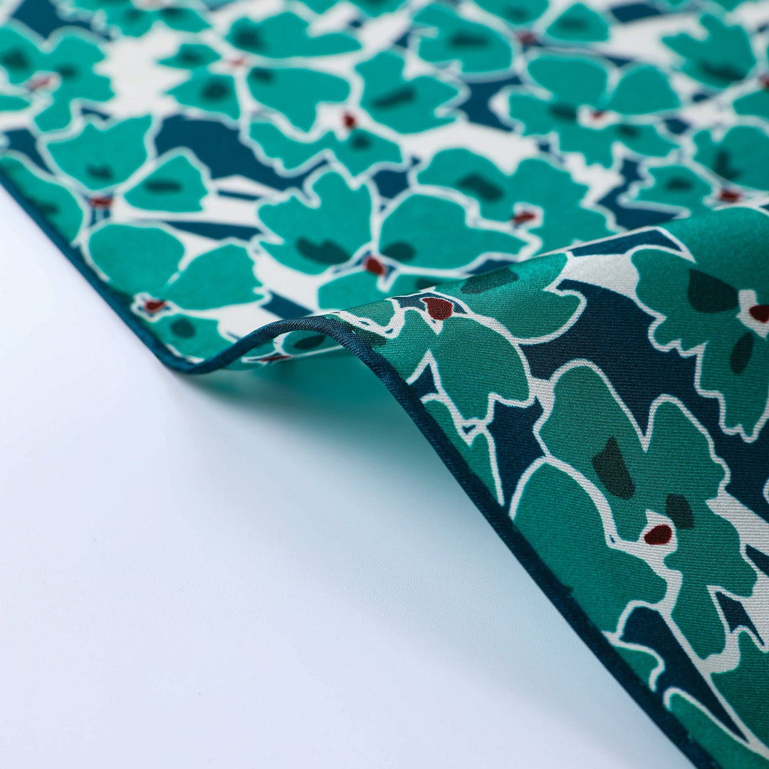 Close-up of the Green & Flourishing Flower Square Silk Scarf, featuring a floral pattern in shades of teal, green, and white with small red accents. This beautiful silk piece from the SusanSilk collection is shown partially folded over on a white surface.