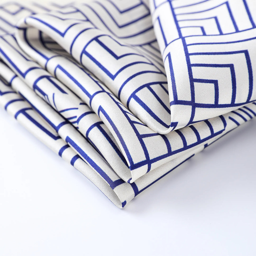 The Land Off Blue Square Silk Scarf by SusanSilk, featuring a geometric blue pattern on white silk fabric, is elegantly displayed on a flat surface, showcasing an exquisite blend of modern design and sophistication.