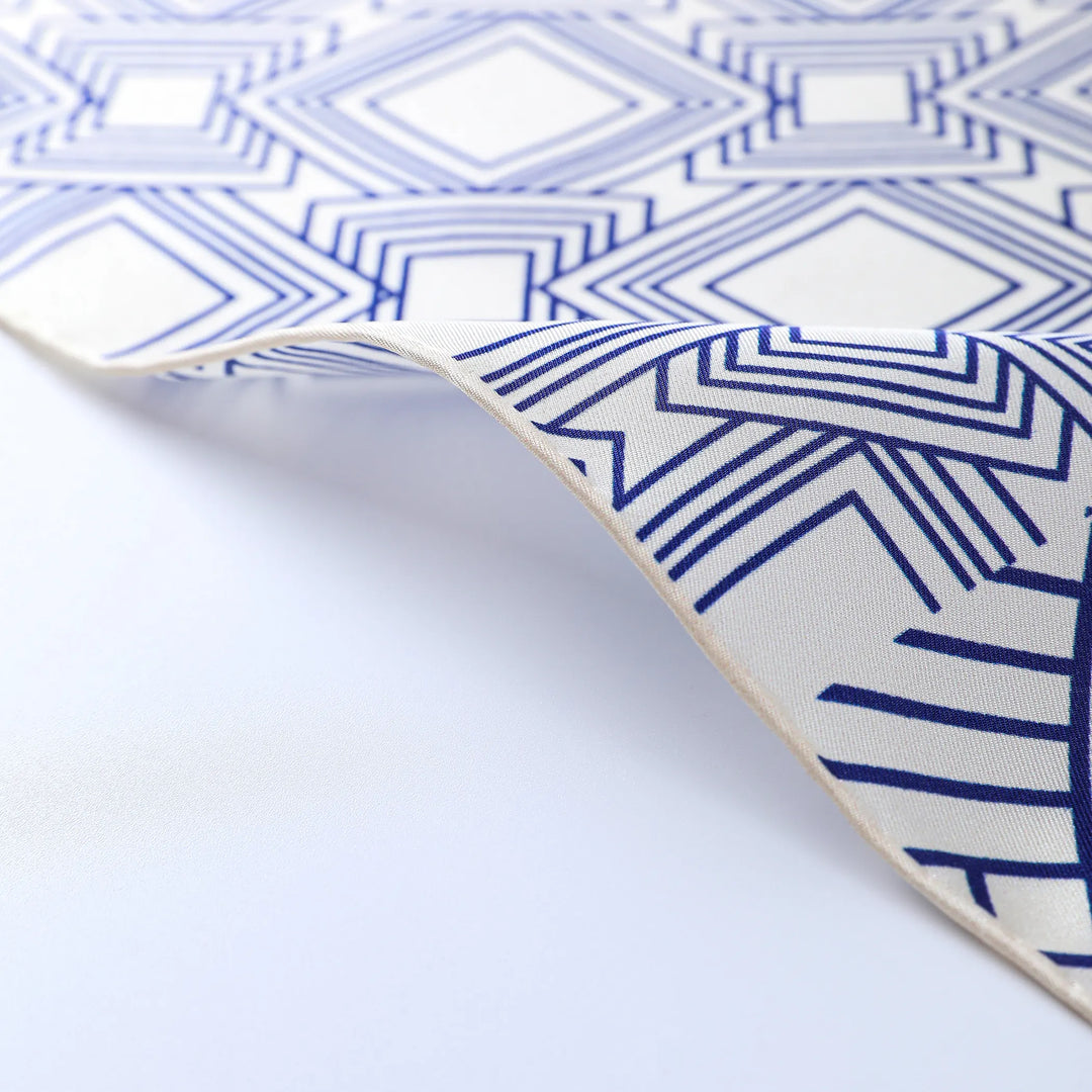 A close-up view of the Land Off Blue Square Silk Scarf from SusanSilk displays a blue and white geometric pattern featuring diamond and linear designs. The scarf is slightly curled at the edge.